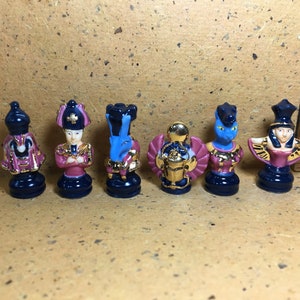 May include: Set of six miniature ceramic chess pieces in an Egyptian theme. The pieces are painted in shades of blue, gold, and pink with intricate details. The pieces include a king, queen, bishop, knight, rook, and pawn.