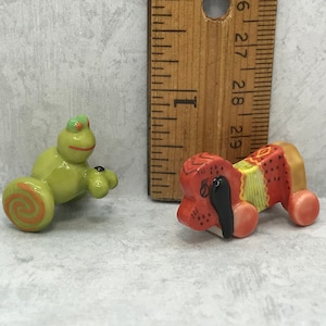 Könnte beinhalten: Zwei kleine Keramikfiguren, ein grüner Frosch mit orangefarbenen Rädern und ein roter und gelber Hund mit braunen Rädern.