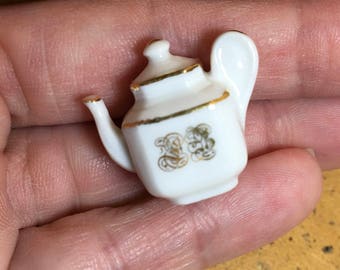 May include: A small white porcelain teapot with a gold trim and a gold design on the front. The teapot has a small spout and a handle.