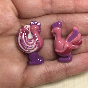 May include: Two ceramic rooster figurines, one pink with purple accents and the other with a white, pink, and purple swirl design. Both roosters have purple feet.