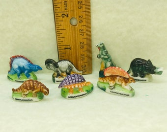Może przedstawiać: Zestaw ośmiu miniaturowych ceramicznych figurek dinozaurów. Każda figurka jest pomalowana żywymi kolorami i przedstawia inny gatunek dinozaura, w tym Stegozaura, Ankylozaura, Scelidozaura, Edaphozaura i wiele innych. Figurki są umieszczone na zielonej podstawie z nadrukowaną nazwą dinozaura.