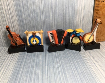 May include: A collection of miniature musical instruments, including a violin, a gong, an accordion, a drum set, and a banjo. Each instrument is set on a black base. The instruments are painted in various colors, including brown, orange, and yellow.