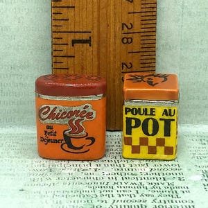 May include: Two miniature rectangular metal tins placed next to a wooden ruler showing a height of approximately 0.75 inches. The left orange tin features the text Chicorée au Petit Déjeuner and a coffee cup. The right yellow tin features the text POULE AU POT.