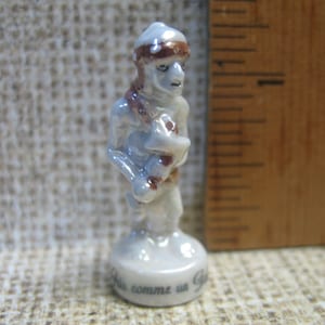 May include: A white porcelain figurine of a person wearing a brown hat and holding a brown object. The figurine is standing on a white base with the text "C'est comme un Gitan" written on it.