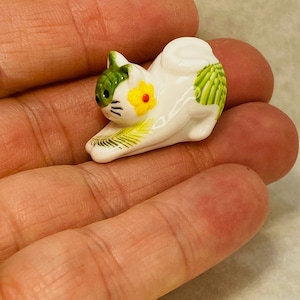 May include: A small white ceramic cat figurine in a stretching pose, featuring green leaf patterns on its head and back, a yellow flower with a red center on its cheek, and black whiskers. A wooden ruler is visible at the top for scale.