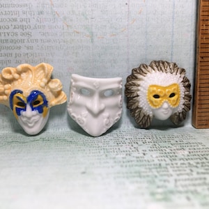 Pode incluir: Três máscaras de carnaval de cerâmica. Uma máscara é branca com um design simples. Uma máscara é amarela com detalhes azuis e amarelos. Uma máscara é branca com detalhes marrons e amarelos.