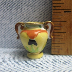 Può includere: Un piccolo vaso in ceramica giallo con accenti dorati e un design dipinto di foglie nere e verdi. Il vaso ha due manici e una base arrotondata.