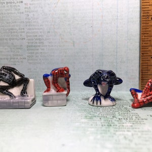 May include: Four ceramic figurines of Spiderman in different poses. One is black and white, one is red and blue, and two are red and black.