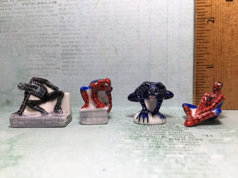 May include: Four ceramic figurines of Spiderman in different poses. One is black and white, one is red and blue, and two are red and black.