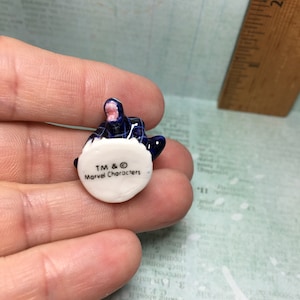 May include: A small, blue and white ceramic figurine of a character from the Marvel universe. The figurine is sitting on a white base with the text "TM & &copy; Marvel Characters" printed on it.