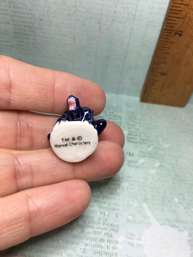 May include: A small, blue and white ceramic figurine of a character from the Marvel universe. The figurine is sitting on a white base with the text "TM & &copy; Marvel Characters" printed on it.