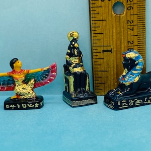 May include: Three miniature Egyptian figurines. The set includes a winged goddess with red, blue, and gold accents, a seated pharaoh, and a sphinx. Each figurine is black with gold detailing and hieroglyphs.