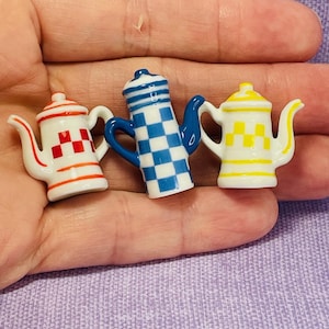 May include: Three miniature ceramic teapots in red, blue, and yellow. The teapots have white bases with checkered and striped patterns. Each teapot has a handle and spout. The teapots are small, likely for a dollhouse or decorative display.