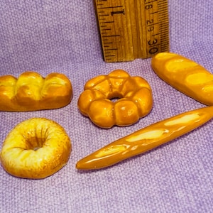May include: Four miniature ceramic bread rolls in various shapes, including a baguette, a round loaf, a braided loaf, and a donut-shaped roll. The rolls are all a light brown colour and are arranged on a purple fabric background.