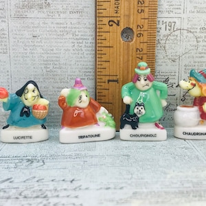 Puede incluir: Cuatro figuras de brujas de cerámica frente a una regla de madera. De izquierda a derecha, las figuras están etiquetadas como Lucifette, Tripatoune, Choupignole y Chaudronasse. Cada figura presenta un atuendo, colores y accesorios únicos, incluyendo una cesta, un gato y un caldero, sobre bases blancas.