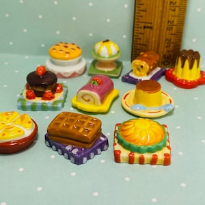 May include: A collection of ten miniature ceramic dessert plates featuring various sweet treats, including a chocolate cake, a yellow cake, a waffle, a fruit tart, a pudding, a roll cake, and a pie. The plates are decorated with colorful patterns and designs.