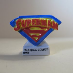 May include: A small, ceramic Superman logo figurine. The logo is a blue diamond shape with a red and yellow "S" emblem and the word "SUPERMAN" in red. The base is white with text.