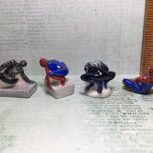 May include: Four small ceramic figurines of Spiderman in different poses. The figurines are painted in red, blue, and black. They are standing on small white bases.
