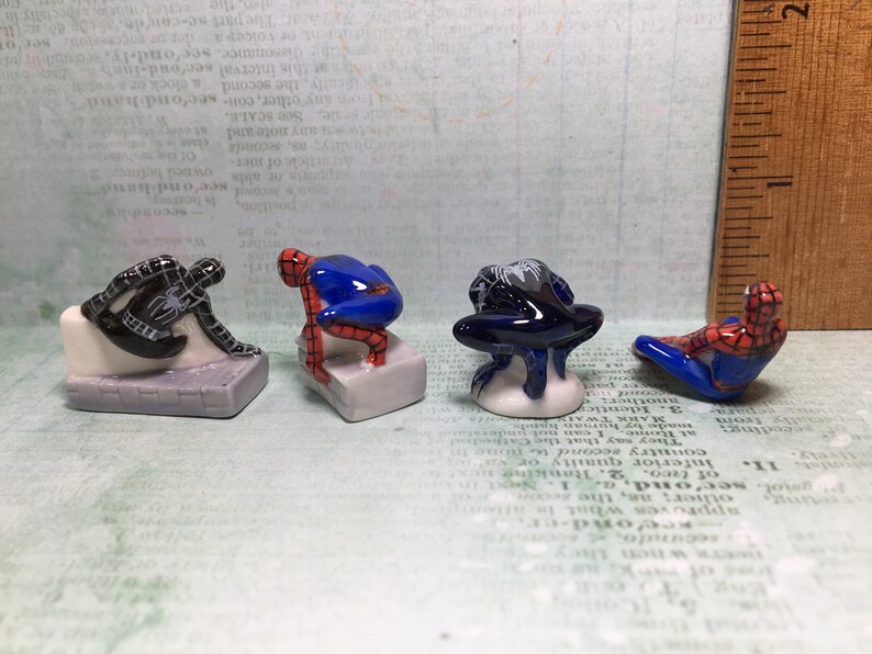 May include: Four small ceramic figurines of Spiderman in different poses. The figurines are painted in red, blue, and black. They are standing on small white bases.