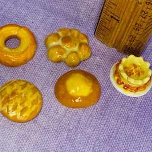 May include: Five miniature ceramic bread rolls, each with a different shape and design. The rolls are yellow and brown and have a glazed finish.