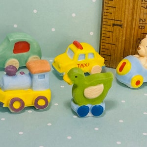 May include: A set of five miniature toy vehicles, including a green car, a yellow taxi, a blue car with a teddy bear driver, a green bird, and a yellow train. The toys are made of ceramic and have painted details.