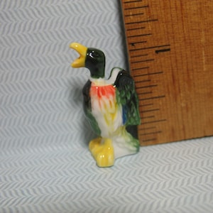 May include: A small ceramic duck figurine with a yellow beak, green and blue feathers, and red and white accents. The duck is standing with its head tilted back and its beak open.