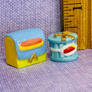 Puede incluir: Dos figuritas de cerámica en miniatura, una una caja azul y amarilla con un asa dorada, la otra una caja de regalo azul con un lazo dorado y una cinta roja.