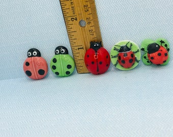 Può includere: Cinque bottoni a forma di coccinella, due rossi con puntini neri, uno verde con puntini neri, uno rosso con puntini neri su una foglia verde e uno rosso con puntini neri su una foglia bianca. I bottoni hanno un diametro di circa 2,5 cm.