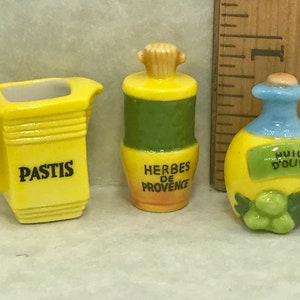 Peut inclure: Trois bouteilles en céramique miniatures avec des couvercles jaunes, verts et bleus. Les bouteilles sont étiquetées "Pastis", "Herbes de Provence" et "Huile d'Olive".