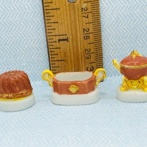 May include: Three miniature porcelain figurines: a brown and gold bundt cake, a brown and gold oval serving dish with handles, and a brown and gold tureen with a lid. The figurines are on a blue background.