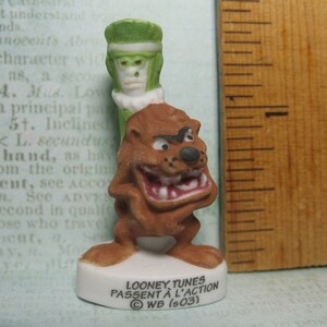 Op de afbeelding: Een keramiek beeldje van een bruine cartoon hond met een groen cartoon personage erop. Het beeldje staat op een witte voet met de tekst "LOONEY TUNES PASSENT À L'ACTION © WB (503)".