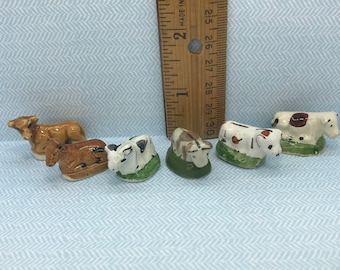 May include: A collection of six miniature ceramic cow figurines. The cows are painted in various colors, including brown, black, and white, and are resting on small green bases. A wooden ruler is in the background.