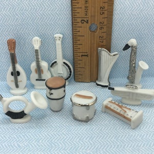 Puede incluir: Colección de instrumentos musicales en miniatura, incluyendo guitarras, un banjo, tambores, una trompeta, un saxofón y un arpa. Los instrumentos son blancos con detalles plateados y marrones, sobre fondo azul claro.