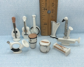 May include: A collection of miniature musical instruments, including guitars, a banjo, drums, a trumpet, a saxophone, and a harp. The instruments are white with silver and brown accents, and are arranged on a light blue patterned surface.