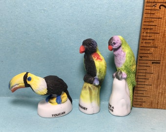 May include: Three miniature ceramic figurines of birds. A toucan with a yellow beak and black and white body, a black and yellow bird with a red beak, and a green and purple bird with a white beak. Each figurine is sitting on a small base with a name written on it: Toucan,  Lorquet, and  Une A Che.