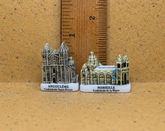 May include: Two miniature ceramic cathedral figurines placed next to a wooden ruler showing a height of approximately 1.5 inches. The left figurine is labeled ANGOULÊME Cathédrale Saint-Pierre, and the right is labeled MARSEILLE Cathédrale de la Major.