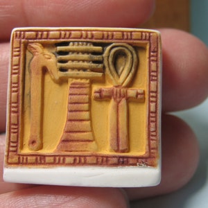 May include: A square-shaped tile featuring a raised relief design in shades of yellow and brown. The central imagery includes an Egyptian-style staff, a djed pillar, and an ankh symbol, all enclosed within a decorative border with etched lines.