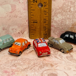 May include: A collection of six miniature ceramic cars in various colors, including blue, orange, red, gray, and black. The cars are all different styles and have varying details, such as a Union Jack flag on the roof of one car.