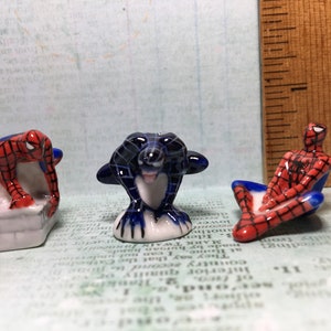 May include: Three ceramic figurines of Spiderman, one in a red and blue suit, one in a black and blue suit, and one in a red and blue suit with a black web design. The figurines are all in different poses.
