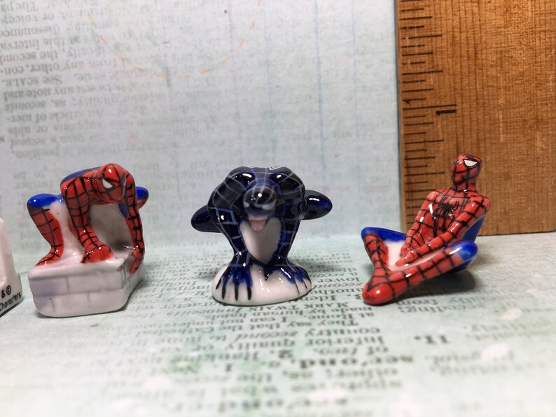 May include: Three ceramic figurines of Spiderman, one in a red and blue suit, one in a black and blue suit, and one in a red and blue suit with a black web design. The figurines are all in different poses.