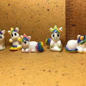 May include: Five small ceramic unicorn figurines with rainbow colored manes and tails. The unicorns are in various poses, including standing, sitting, and lying down.