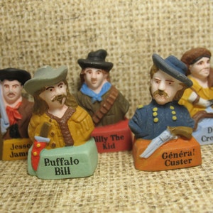 Może przedstawiać: Zestaw sześciu miniaturowych figurek znanych postaci historycznych, każda z kapeluszem i bronią. Figurki są pomalowane realistycznymi kolorami i mają szczegółowe cechy. Nazwy postaci są nadrukowane na podstawach: Jesse James, Buffalo Bill, Billy the Kid, Generał Custer, Davy Crockett.