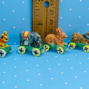 Könnte beinhalten: Vier Miniatur-Tierfiguren aus Keramik auf Rädern. Ein Tiger, ein Elefant, ein Kaninchen und eine Katze sind in leuchtenden Farben bemalt und fahren auf grünen Spielzeugautos mit gelben Rädern.