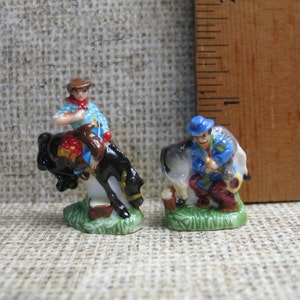 May include: Two miniature ceramic figurines depicting cowboys on horseback. The cowboy on the left is wearing a blue shirt and a brown hat, while the cowboy on the right is wearing a blue hat and a red shirt. Both figurines are standing on a green base.