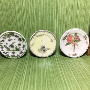 May include: Three miniature ceramic plates with different floral designs. The first plate has a green and white design with a bird and palm trees. The second plate has a yellow and white design with a floral pattern. The third plate has a white and pink design with a bouquet of flowers.