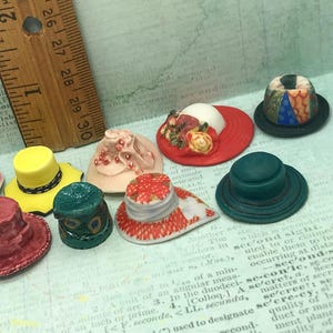 May include: A collection of miniature hats in various colors and styles, including a red hat with floral accents, a yellow hat, a dark green hat, and a patterned hat, positioned next to a wooden ruler showing a measurement of one inch.