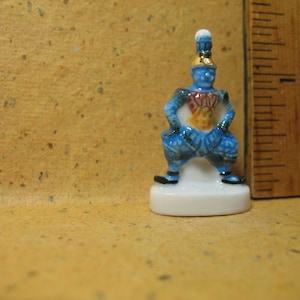 Puede incluir: Una figurita de porcelana azul y roja de un personaje que lleva un sombrero azul y dorado y está sentado sobre una base blanca.