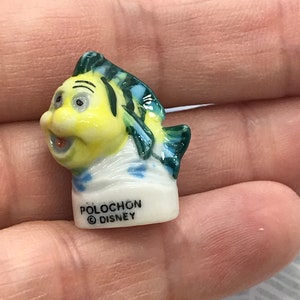 May include: A small ceramic figurine of a yellow fish with green and blue fins. The fish has a smiling face and is wearing a white sailor hat. The figurine is labeled "Polochon &copy; Disney".