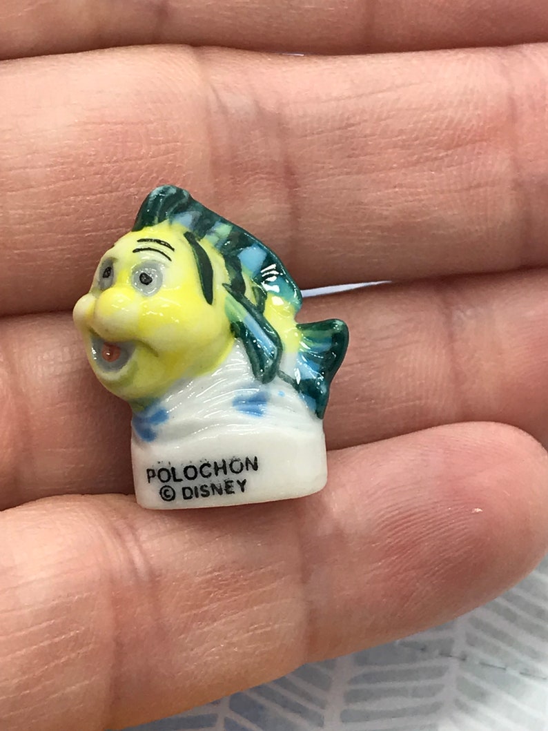 May include: A small ceramic figurine of a yellow fish with green and blue fins. The fish has a smiling face and is wearing a white sailor hat. The figurine is labeled "Polochon &copy; Disney".