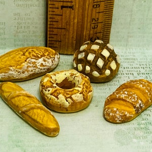 Puede incluir: Una colección de cinco piezas de pan de cerámica en miniatura, que incluyen una baguette, una hogaza redonda, una hogaza larga, una dona y una hogaza redonda con un patrón de diamante. Todos los artículos son de color marrón claro y están dispuestos sobre una superficie texturizada.